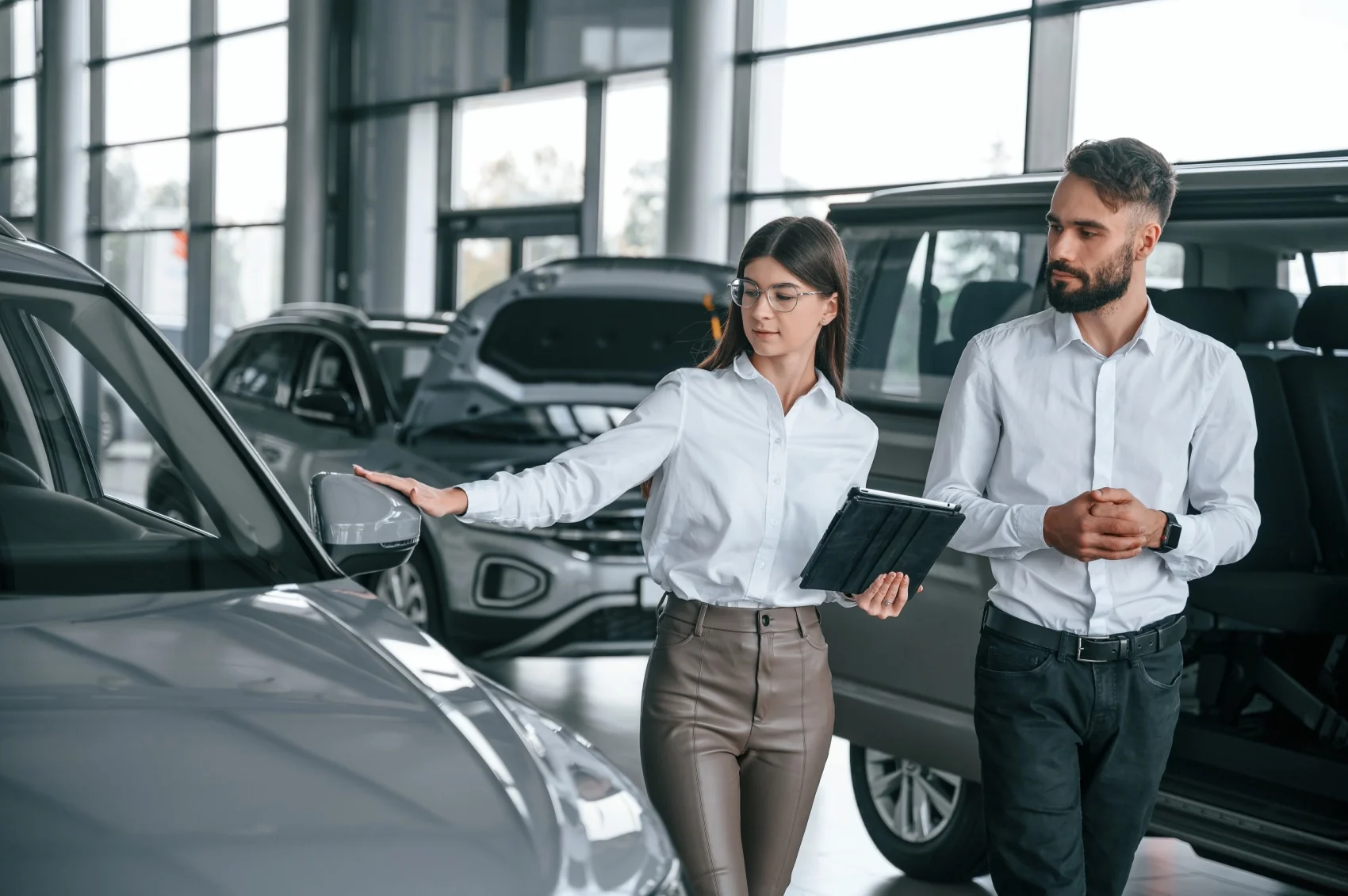 A woman in a white shirt showing a car feature to a man in a white shirt, both in a car dealership surrounded by various vehicles.